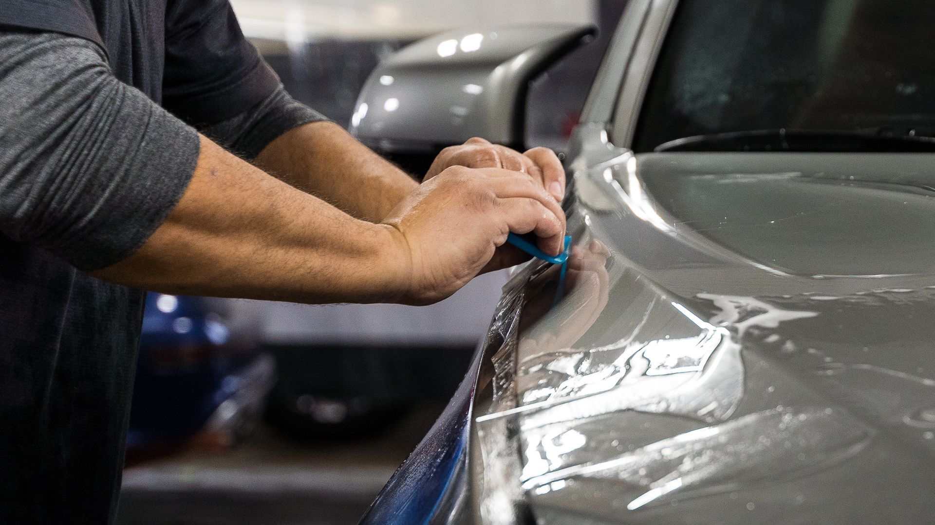 A person applying a protective film to the front of a gray car.