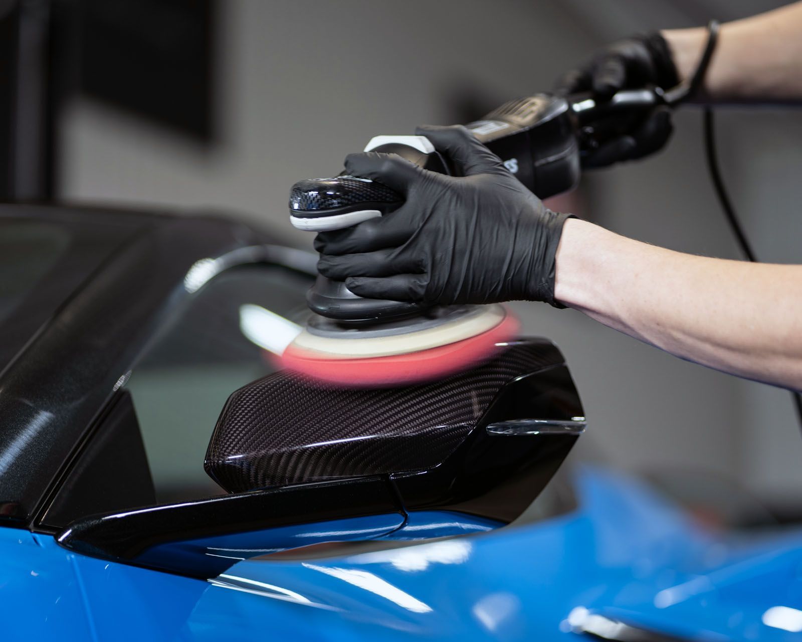 Person in black gloves polishing a blue car's black side mirror with a rotary polisher.
