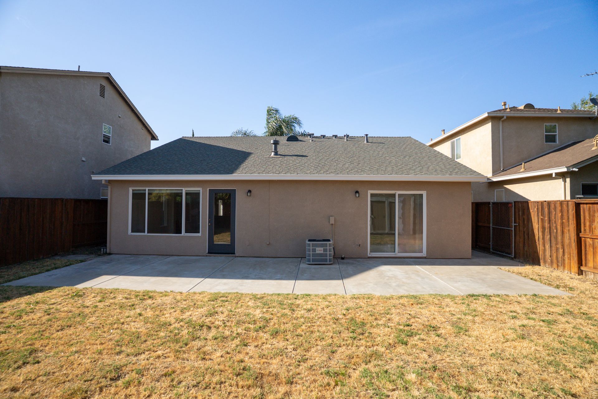 Backyard of a house with a concrete patio and dry grass, flanked by fences and neighboring houses.