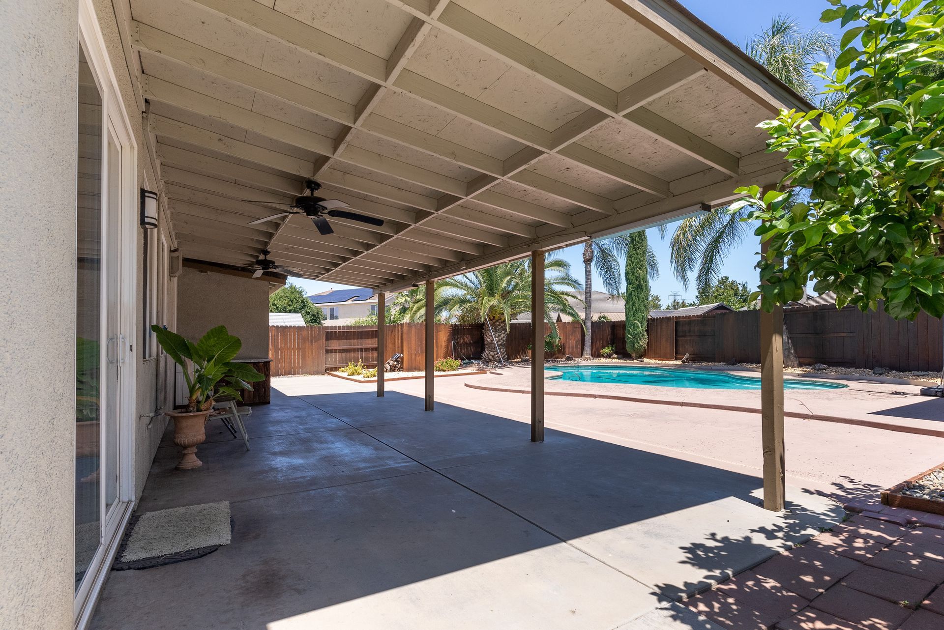 Covered patio overlooking a swimming pool, with brown support beams and a light blue sky.