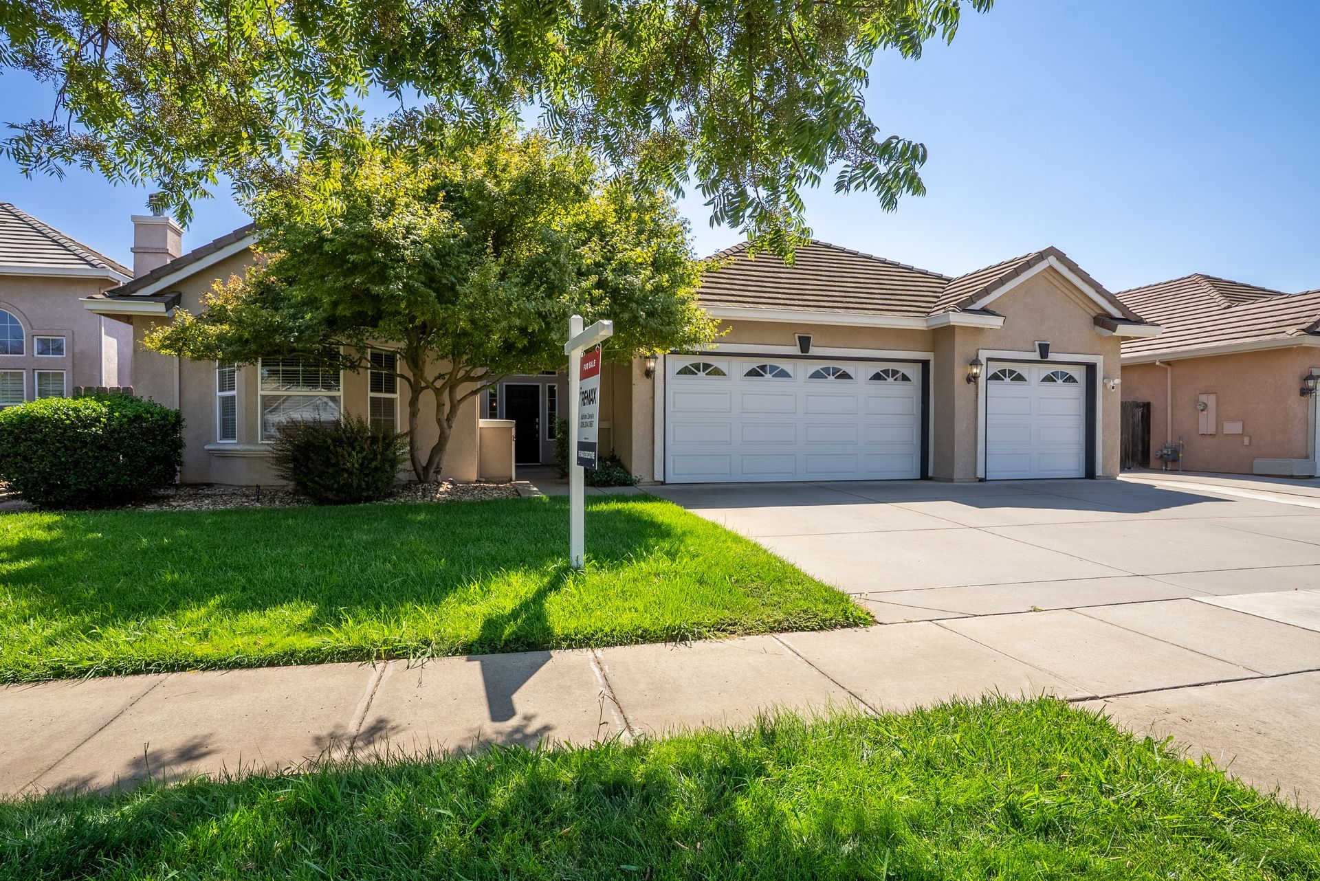 Tan suburban house with a three-car garage, green lawn, and tree in front.