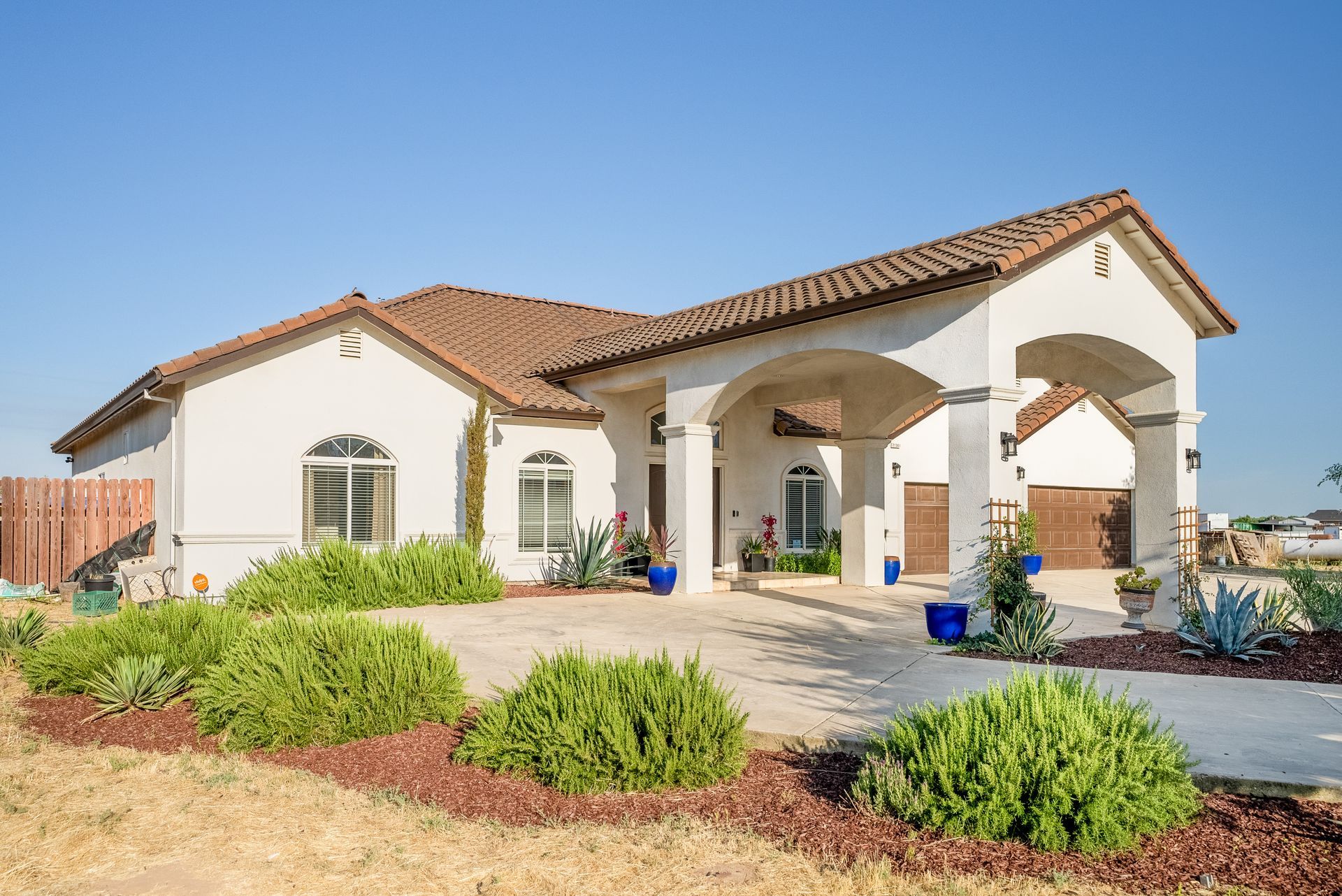 White stucco house with arched entryway and brown tile roof, green bushes in front.