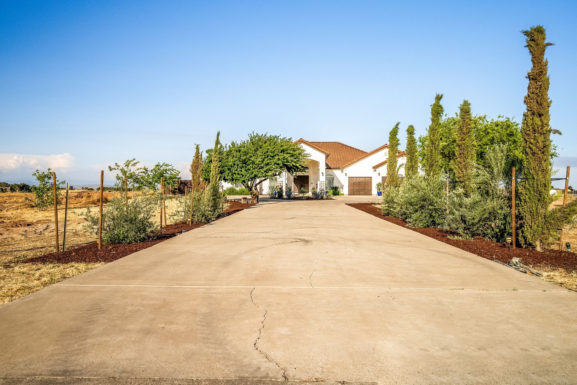 Concrete driveway leading to a white house with landscaping under a blue sky.