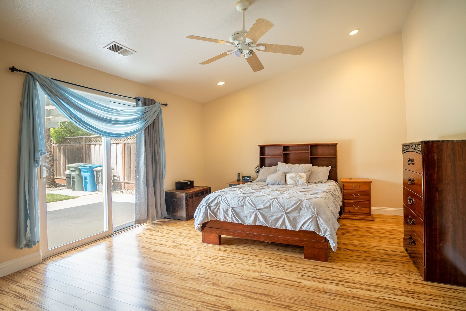 Bedroom with bed, dresser, nightstand, and sliding door. Pale yellow walls, wood floors, and a ceiling fan.