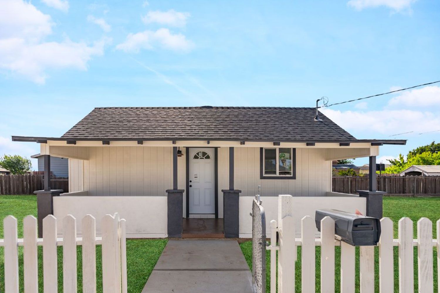 Small, white cottage with a picket fence and mailbox. Blue sky.