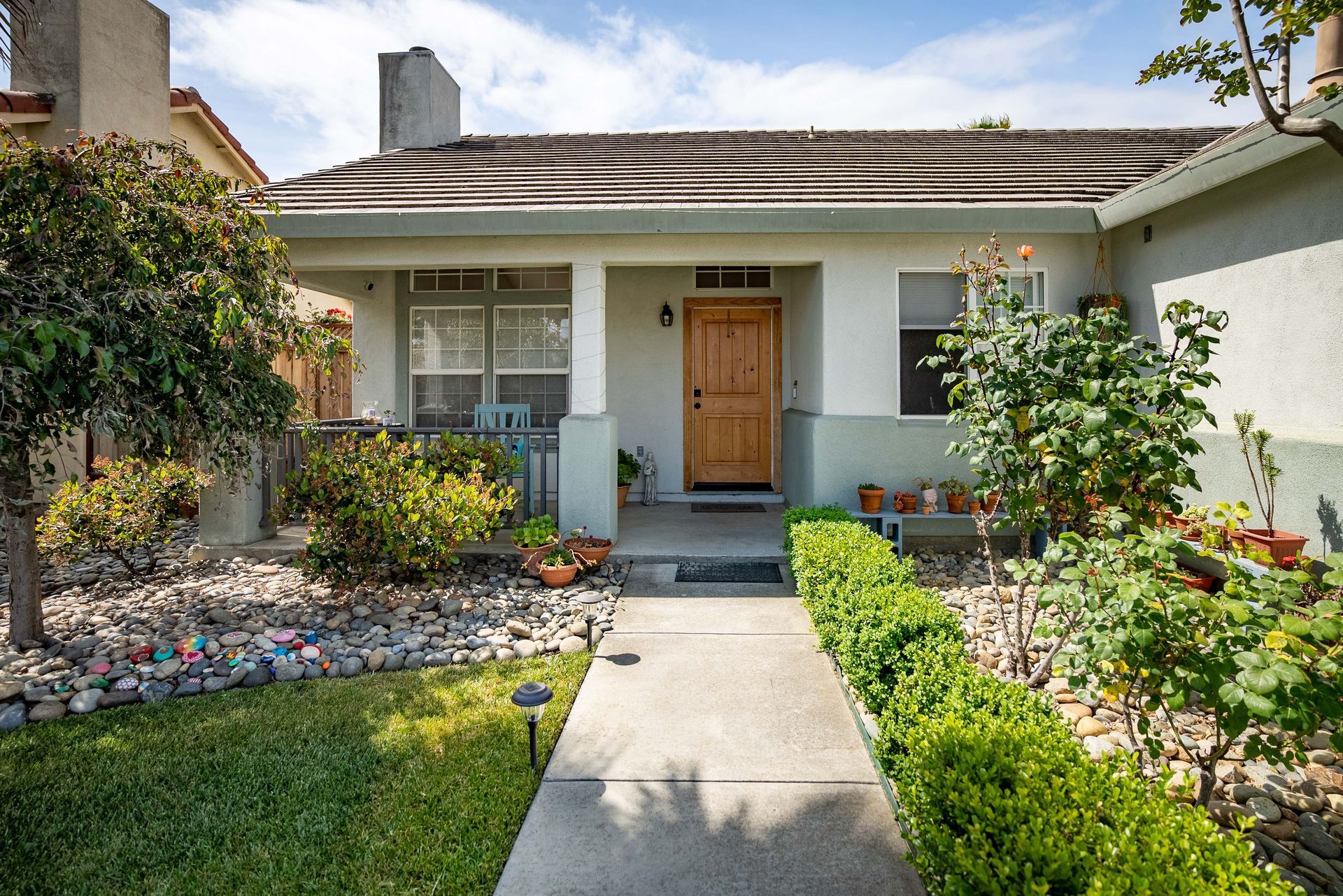 A light-colored house with a covered porch and path leading to a wooden door, surrounded by greenery and a rock garden.