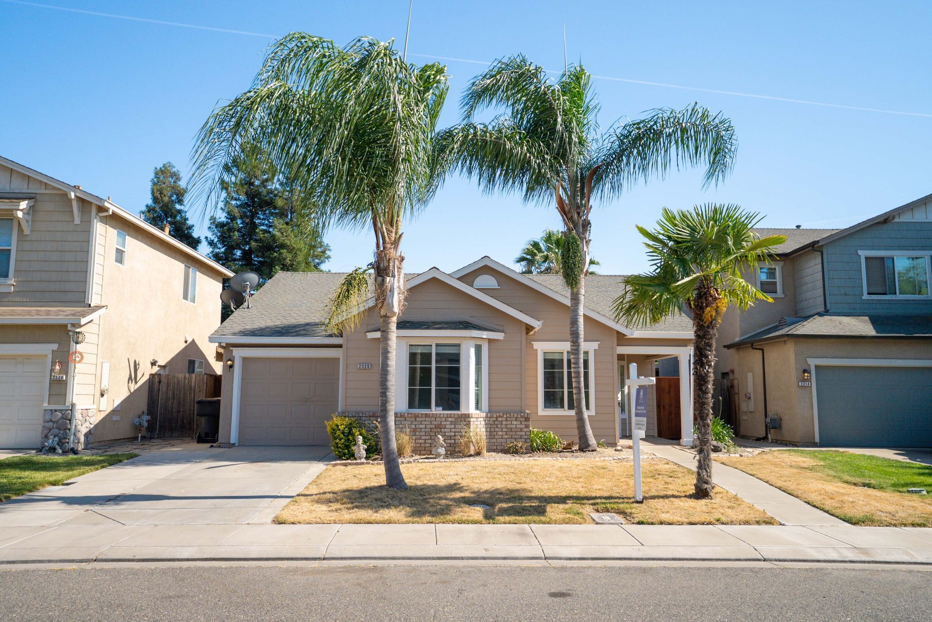 Tan house with palm trees and a for sale sign.