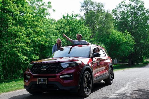 Red SUV on a forest road with two people standing through the sunroof holding arms up