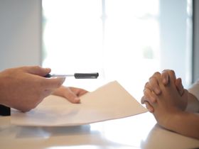 Hands clasped at a desk as a pen points toward paperwork in a bright office setting