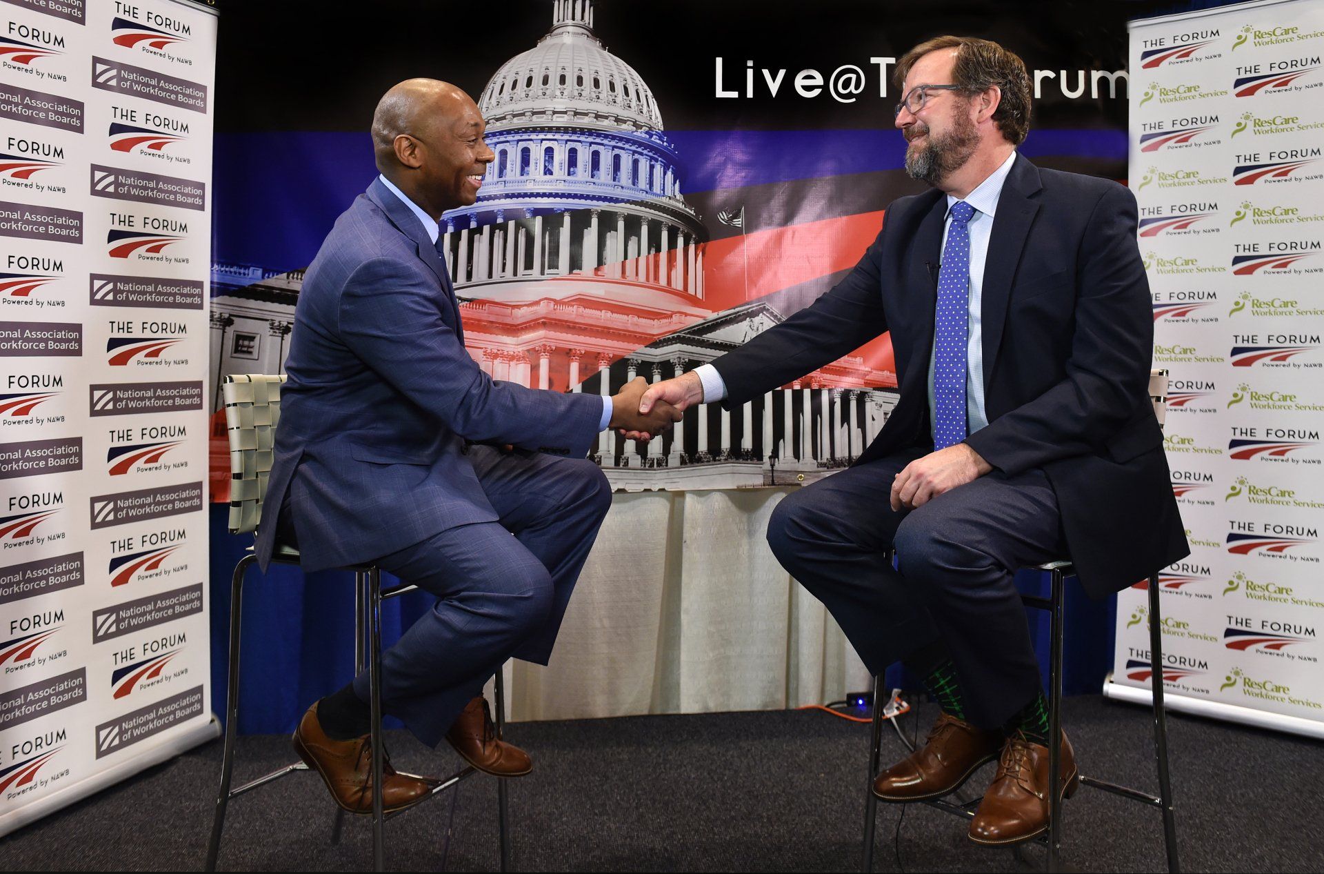 Two men in suits are sitting on stools talking to each other