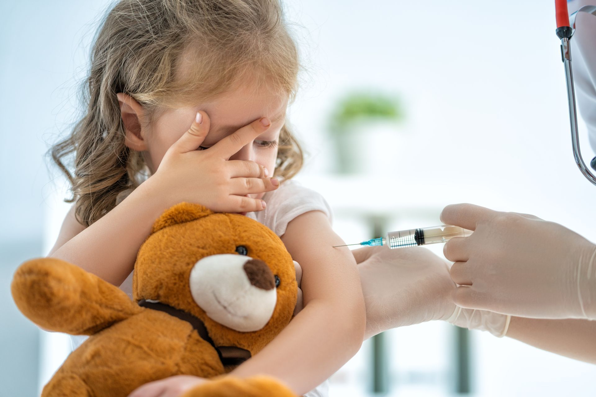 A little girl is holding a teddy bear while getting an injection.