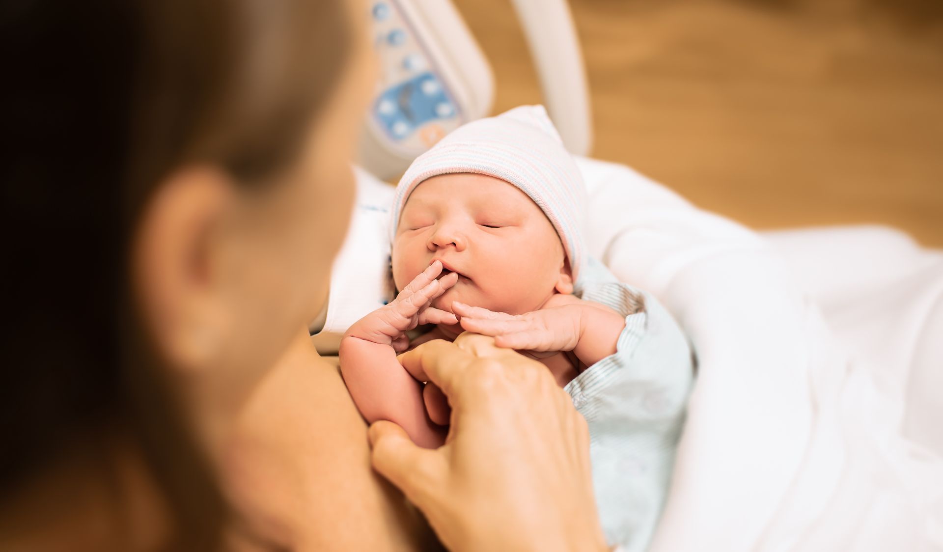 Newborn baby in a white blanket, wearing a hat, held by a person.