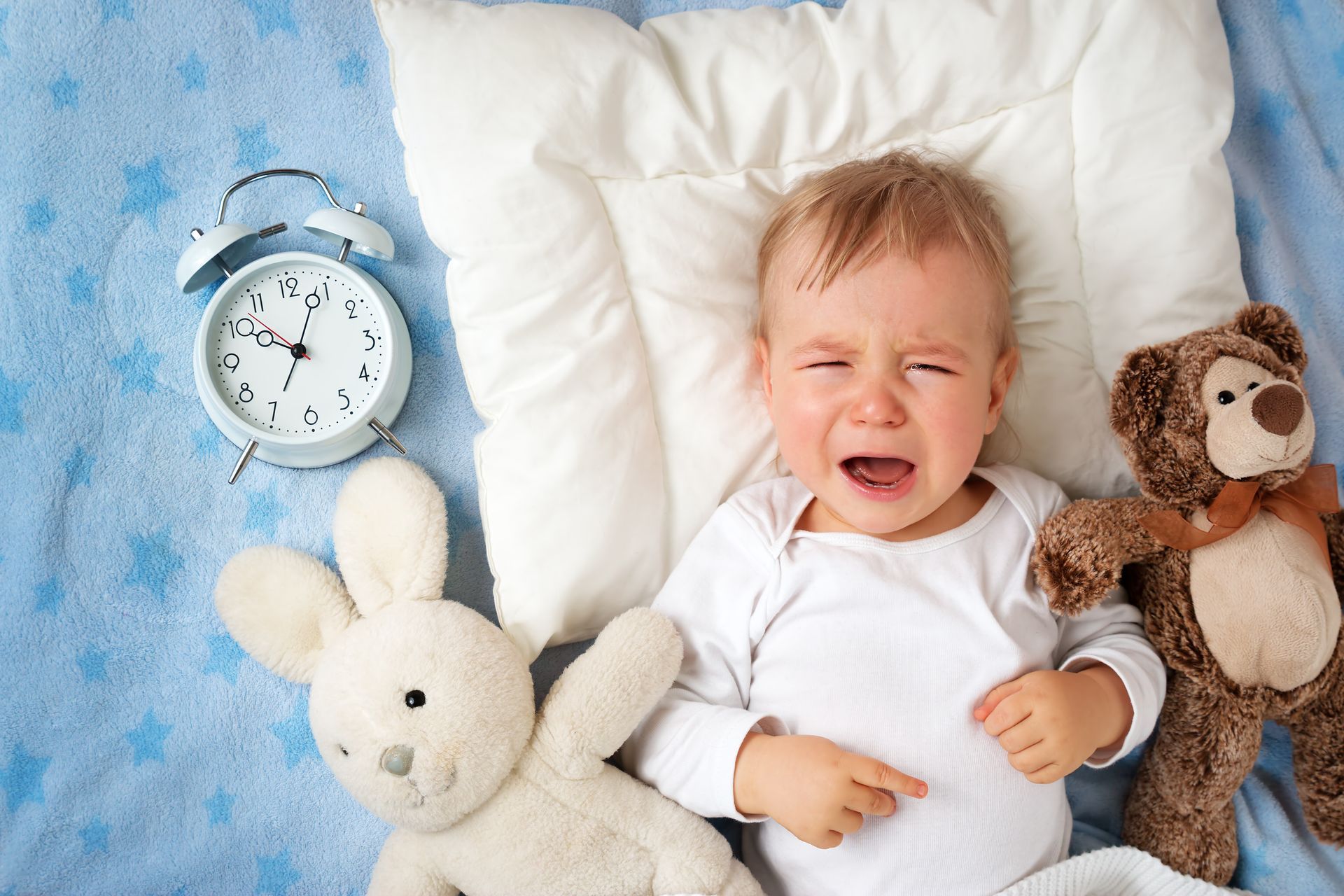 A baby is crying while laying in bed with stuffed animals and an alarm clock.