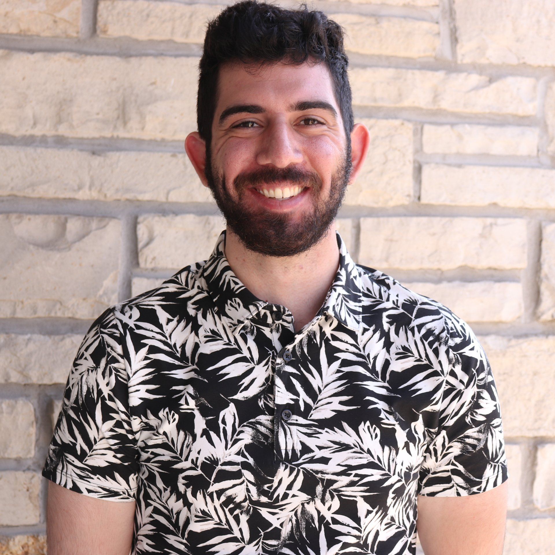 A man with a beard wearing a black and white shirt is smiling in front of a brick wall.