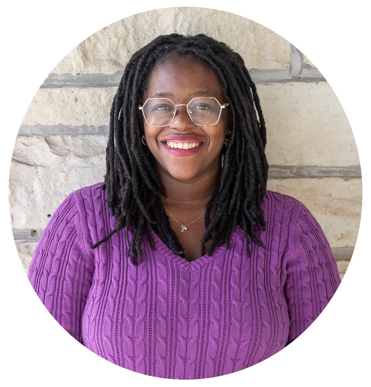 A woman wearing glasses and a purple shirt is smiling in front of a brick wall.