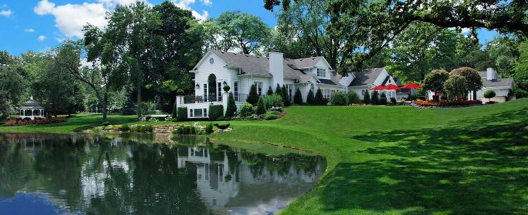 A large white house is sitting next to a lake surrounded by trees.