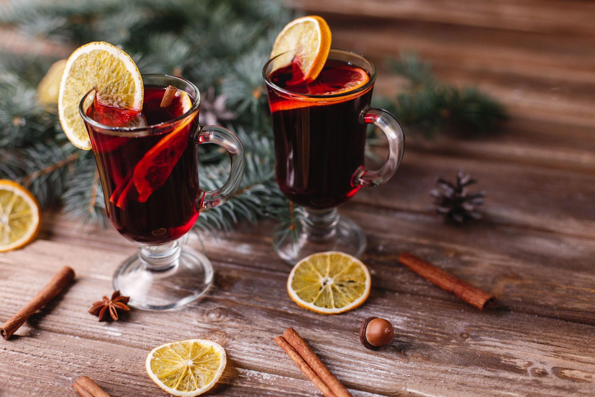 Red cocktails in glass mugs with orange slices and cinnamon sticks on a wood table with pine tree branches.