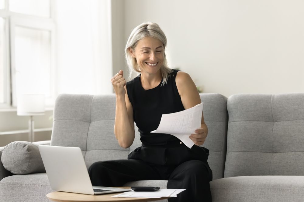 Woman on couch celebrates with paperwork, near laptop and table.