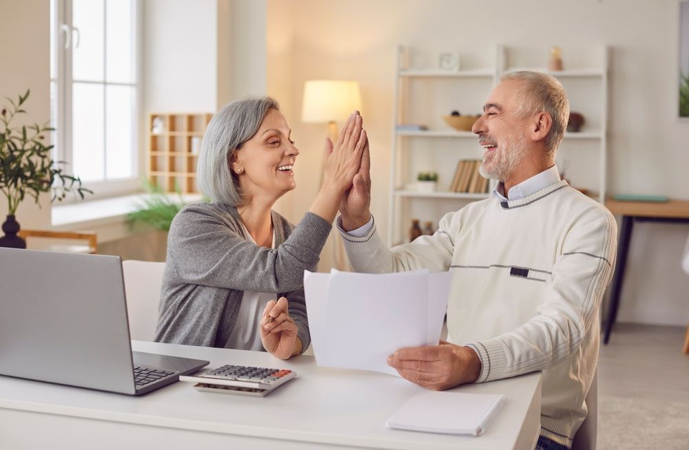 Senior couple high-fiving at a desk with laptop, calculator, and papers, looking happy in a well-lit room.