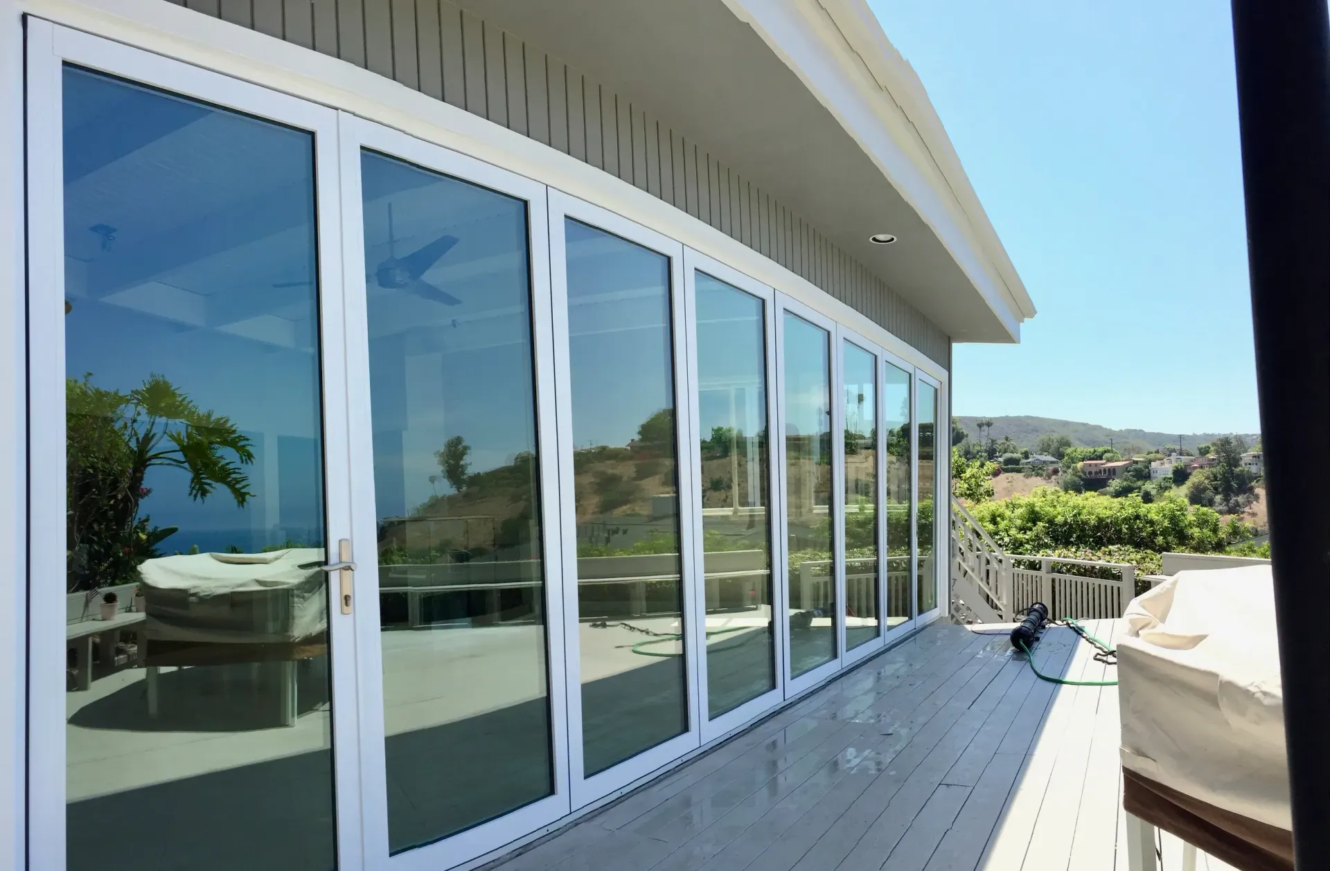 A row of closed, white-framed glass doors reflecting a sunny outdoor scene. Building exterior with gray siding and a wooden deck.