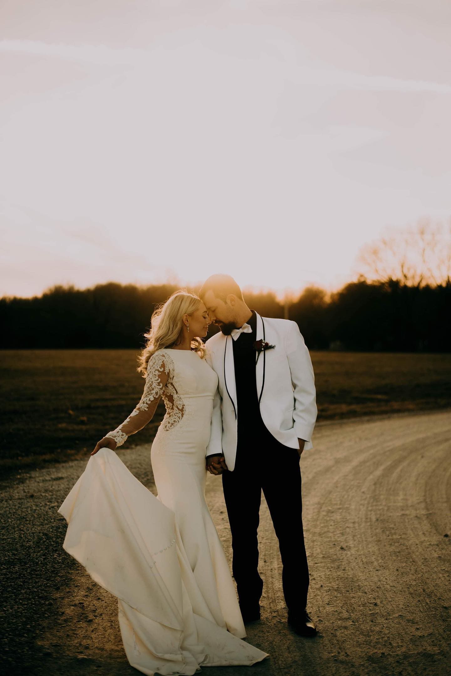 A bride and groom are kissing on a dirt road at sunset.