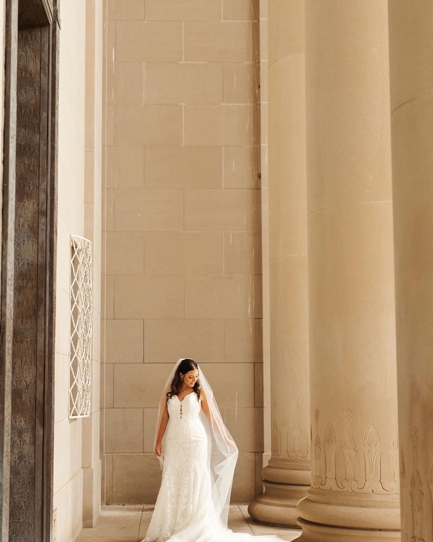 A bride in a wedding dress and veil is standing in front of a building with columns.