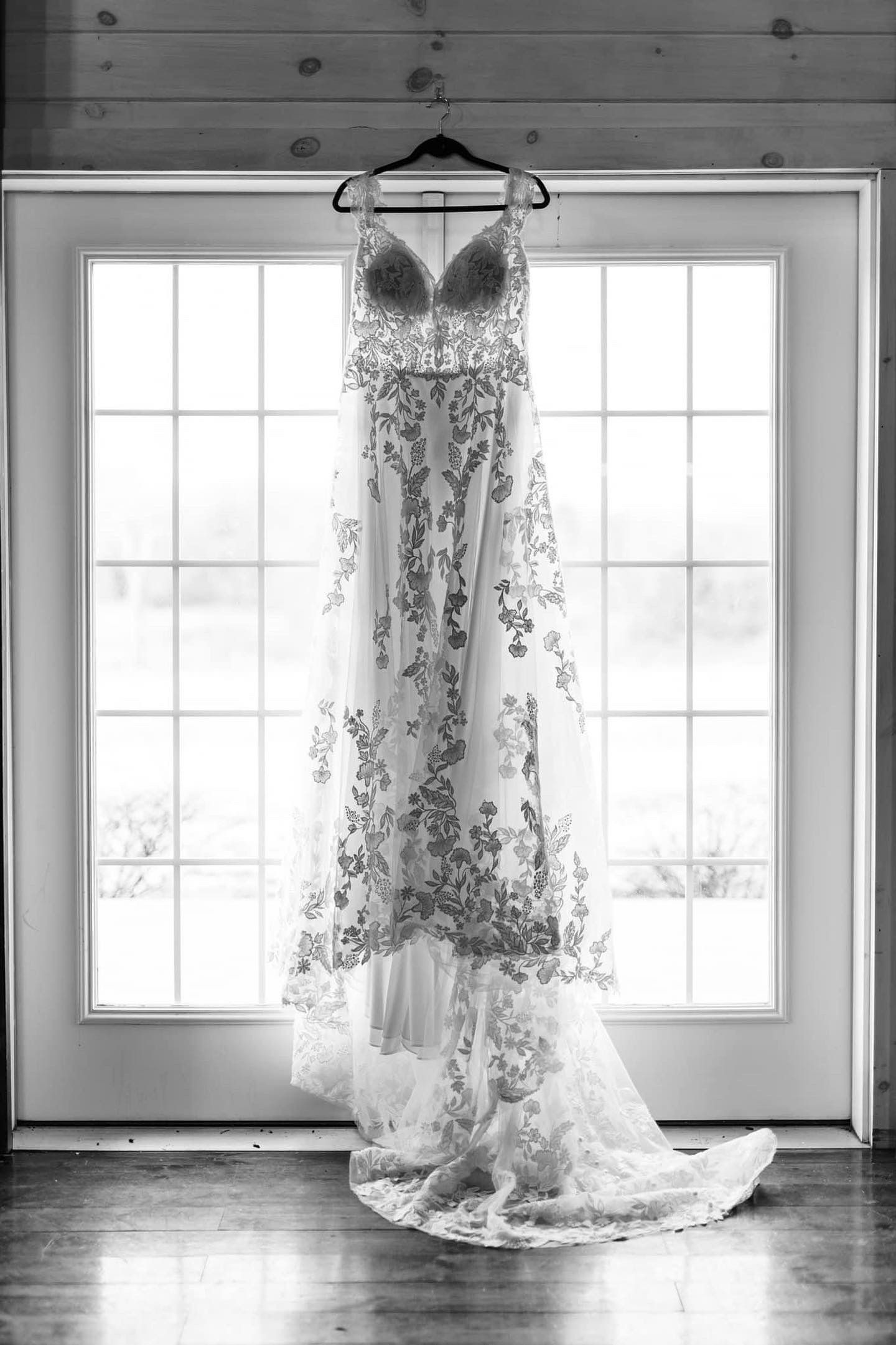 A black and white photo of a wedding dress hanging in front of a window.