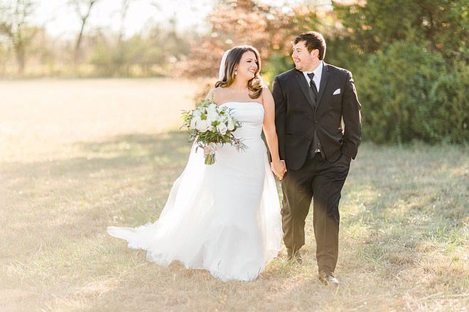 A bride and groom are walking in a field holding hands.
