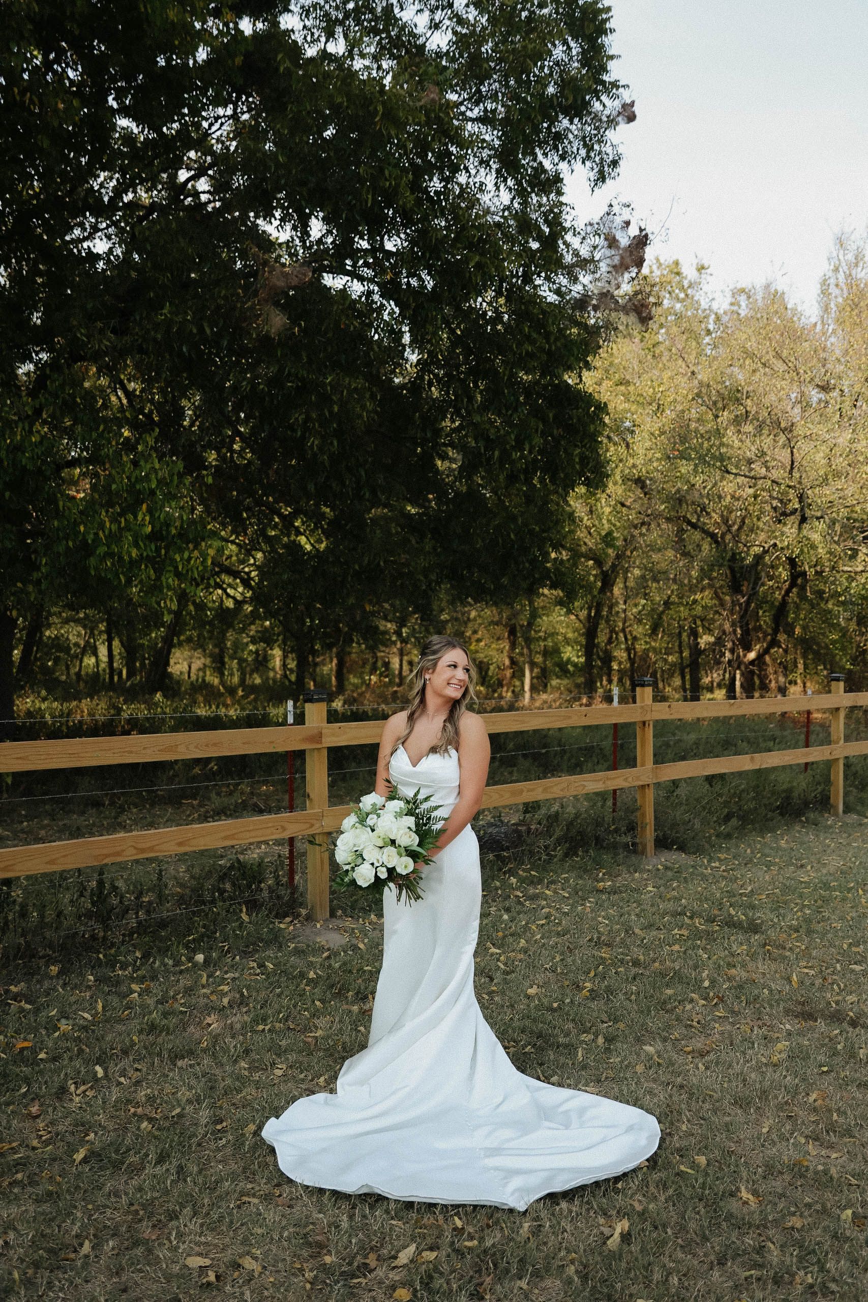 A bride in a wedding dress is standing in front of a wooden fence holding a bouquet of flowers.