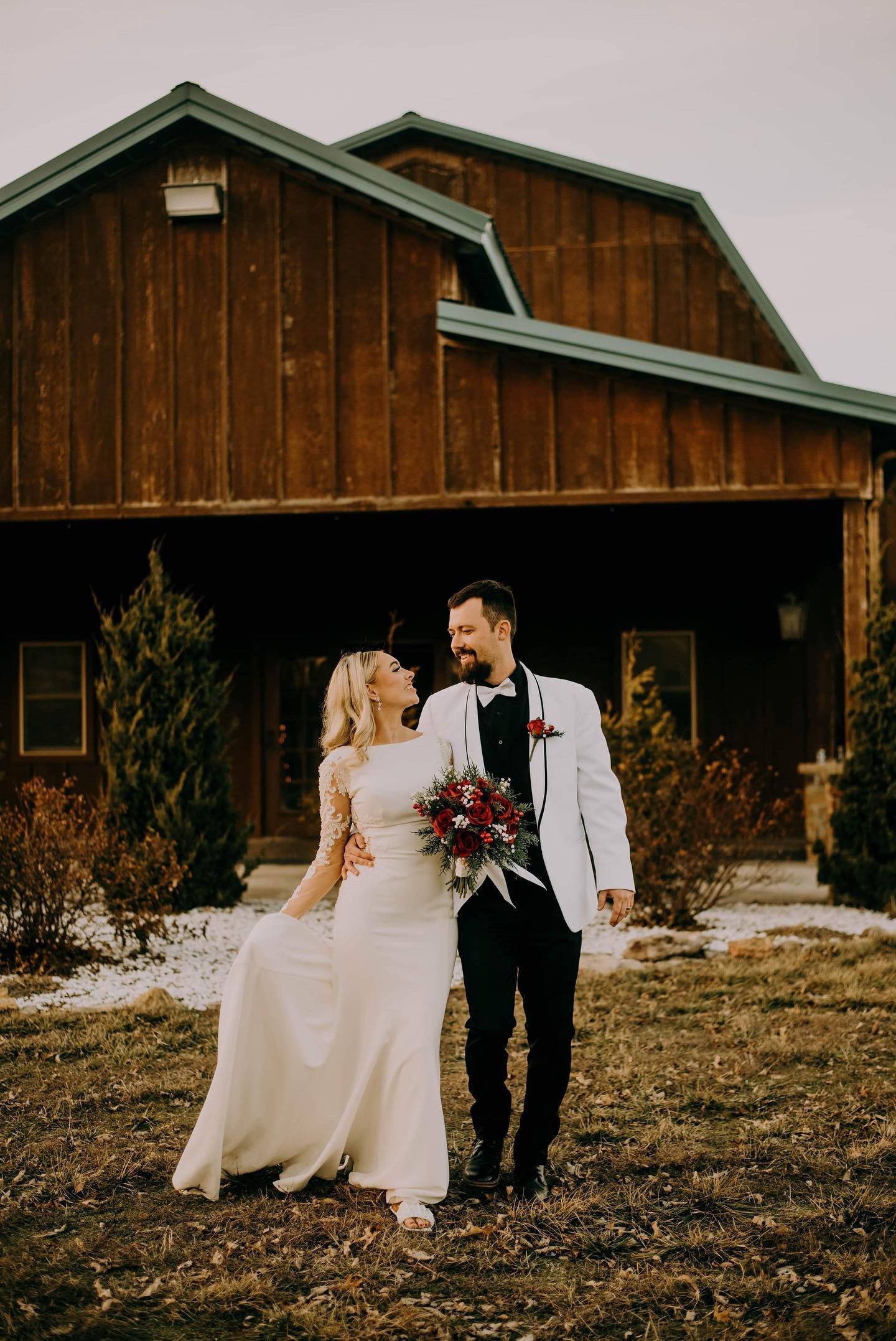 A bride and groom are walking in front of a wooden barn.