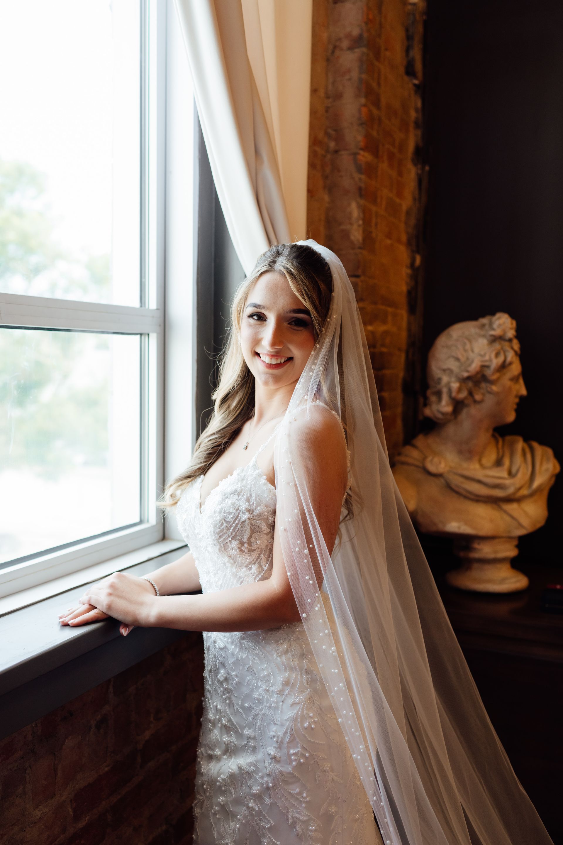 A bride in a wedding dress and veil is standing next to a window.