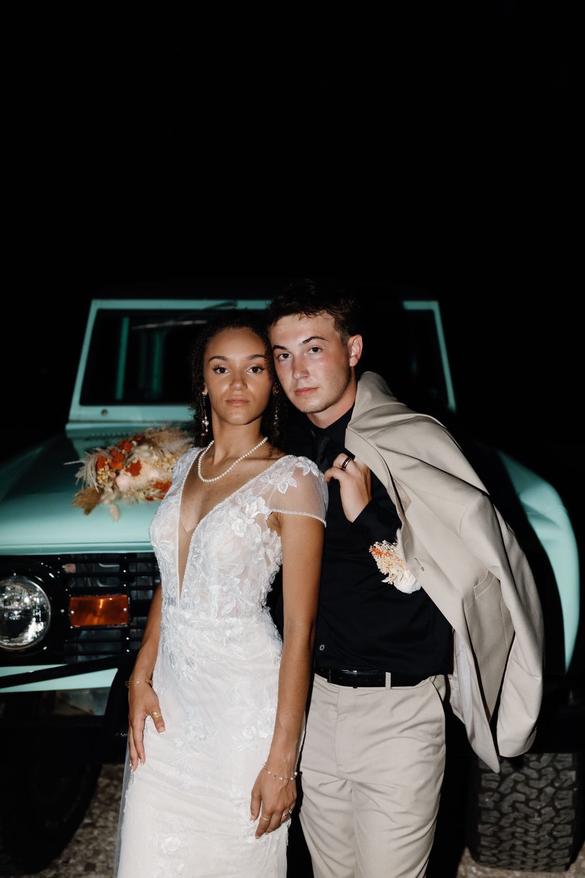 A bride and groom are posing for a picture in front of a car.