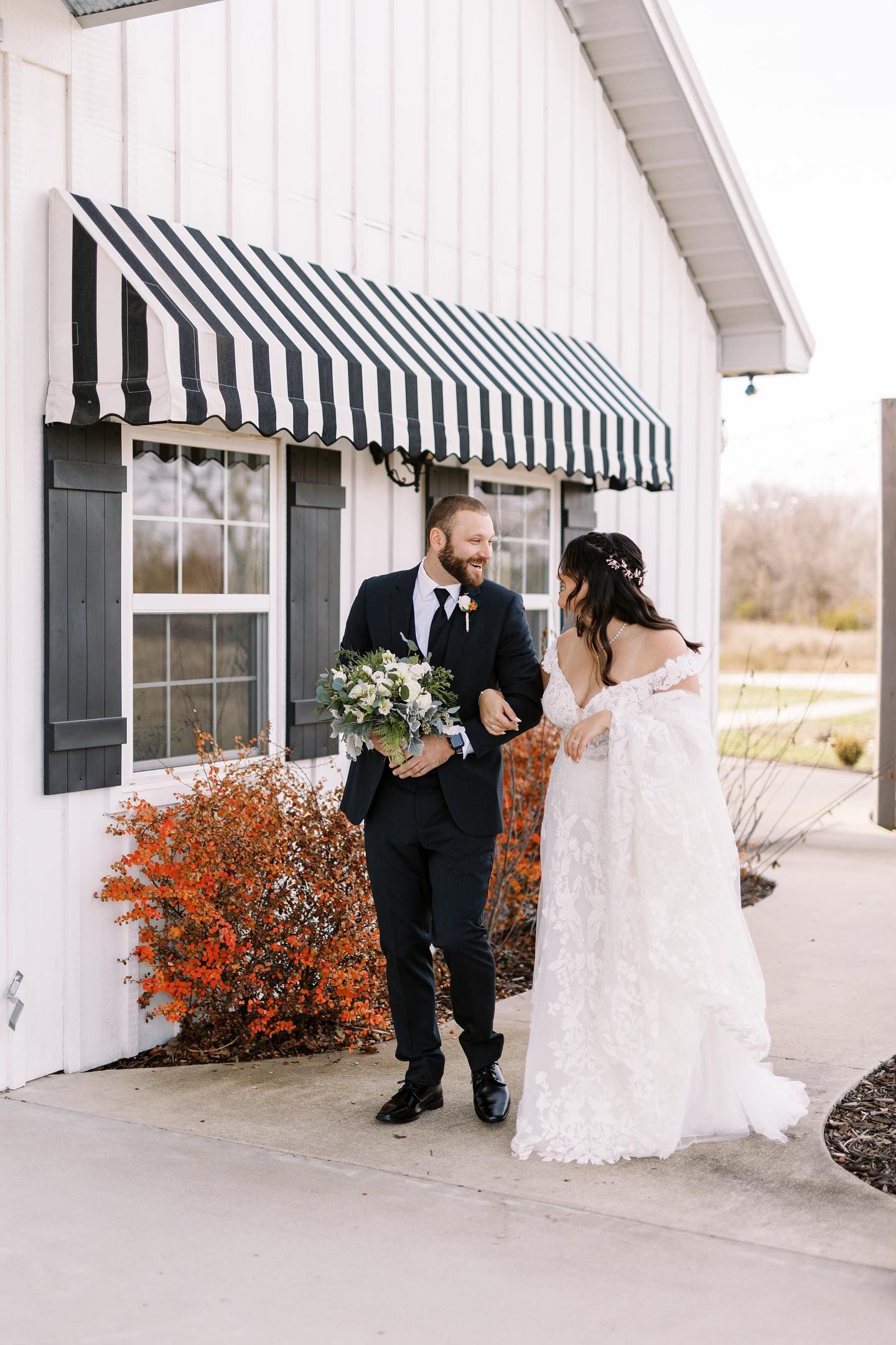 A bride and groom are walking in front of a white building . the bride is holding a bouquet of flowers.