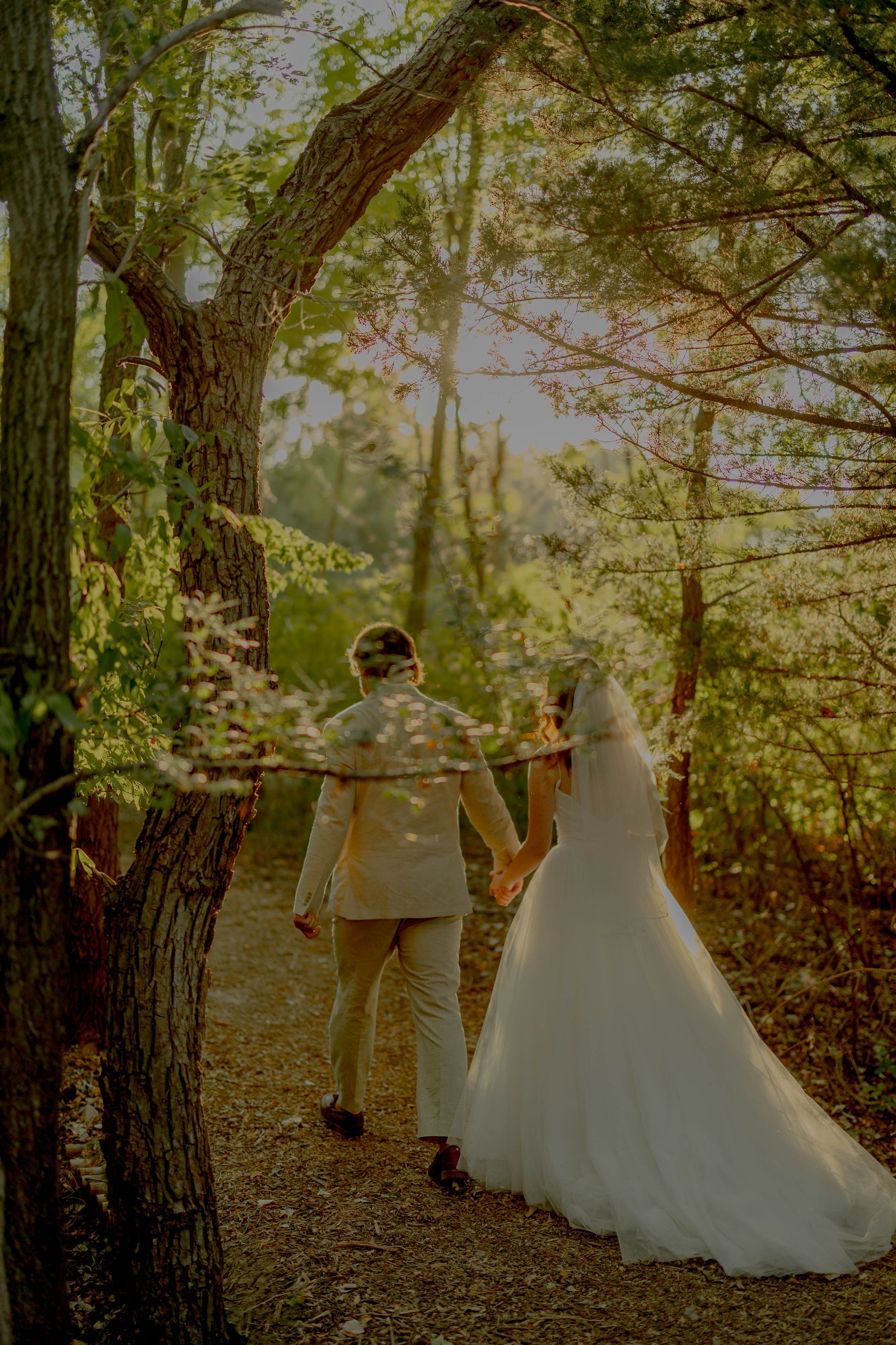 A bride and groom are walking through the woods holding hands.