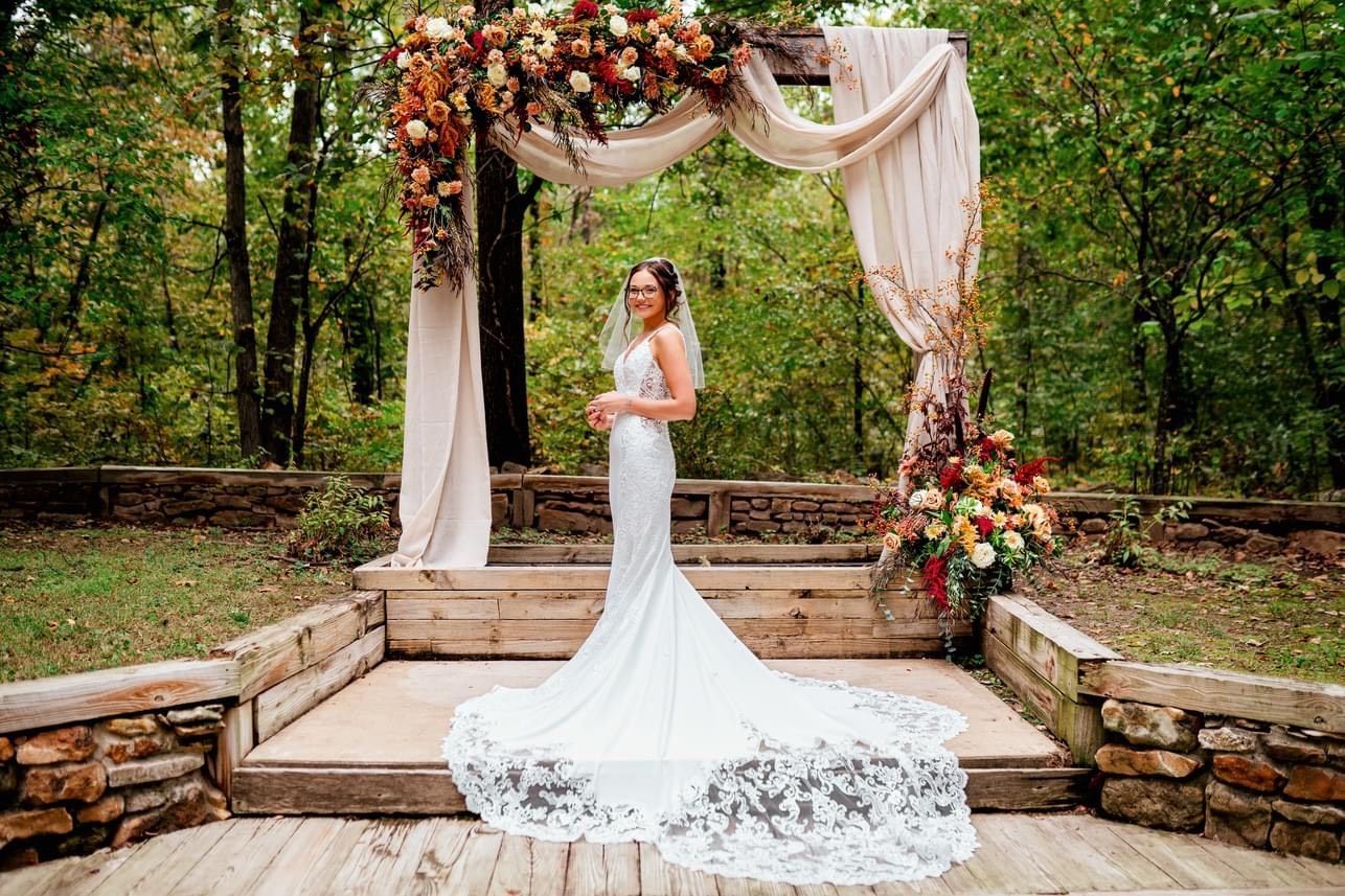 A bride in a wedding dress is standing in front of a wooden arch decorated with flowers.
