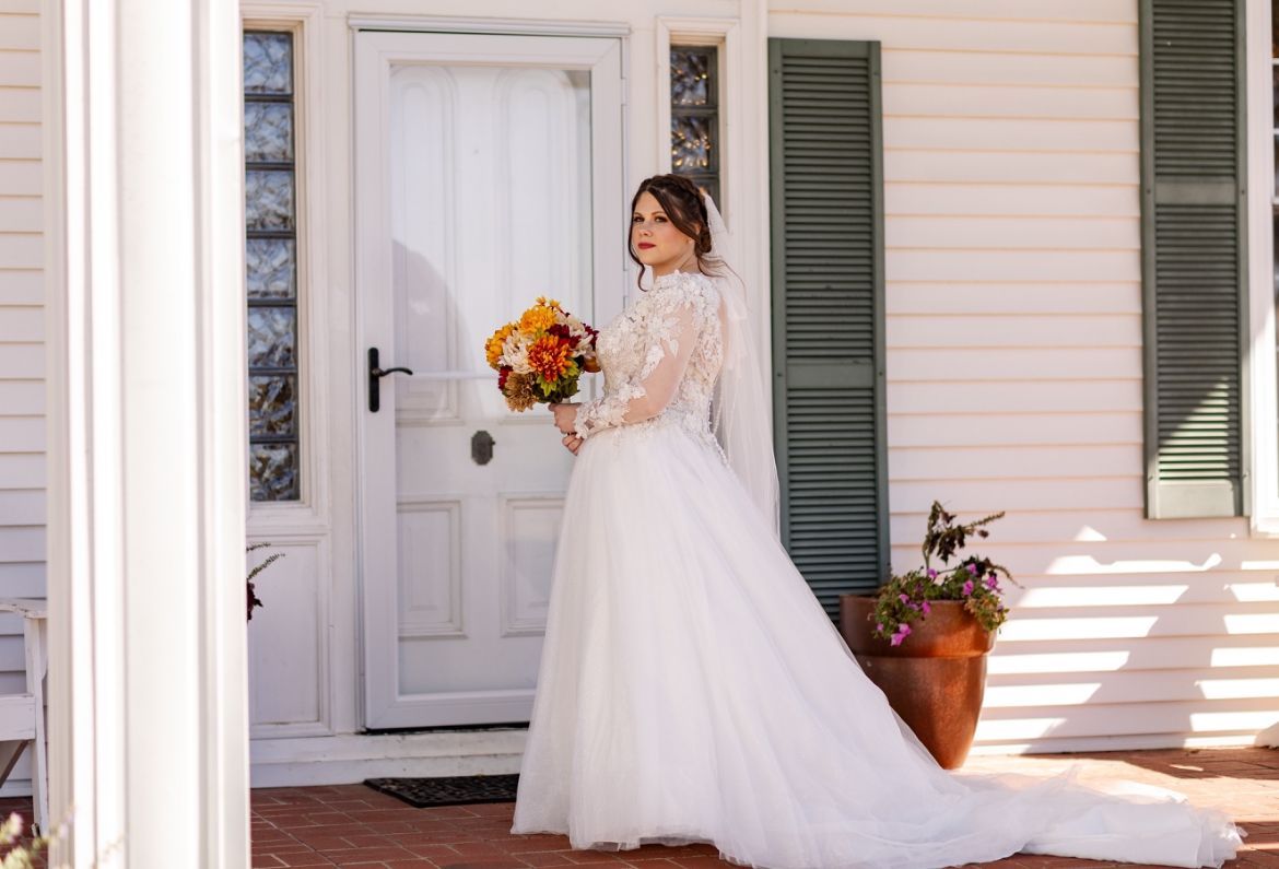 A bride in a wedding dress is standing in front of a white house holding a bouquet of flowers.