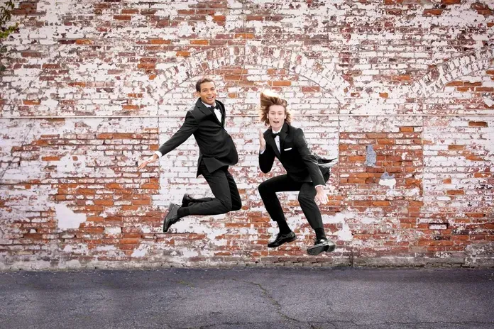 Two men in tuxedos are jumping in the air in front of a brick wall.