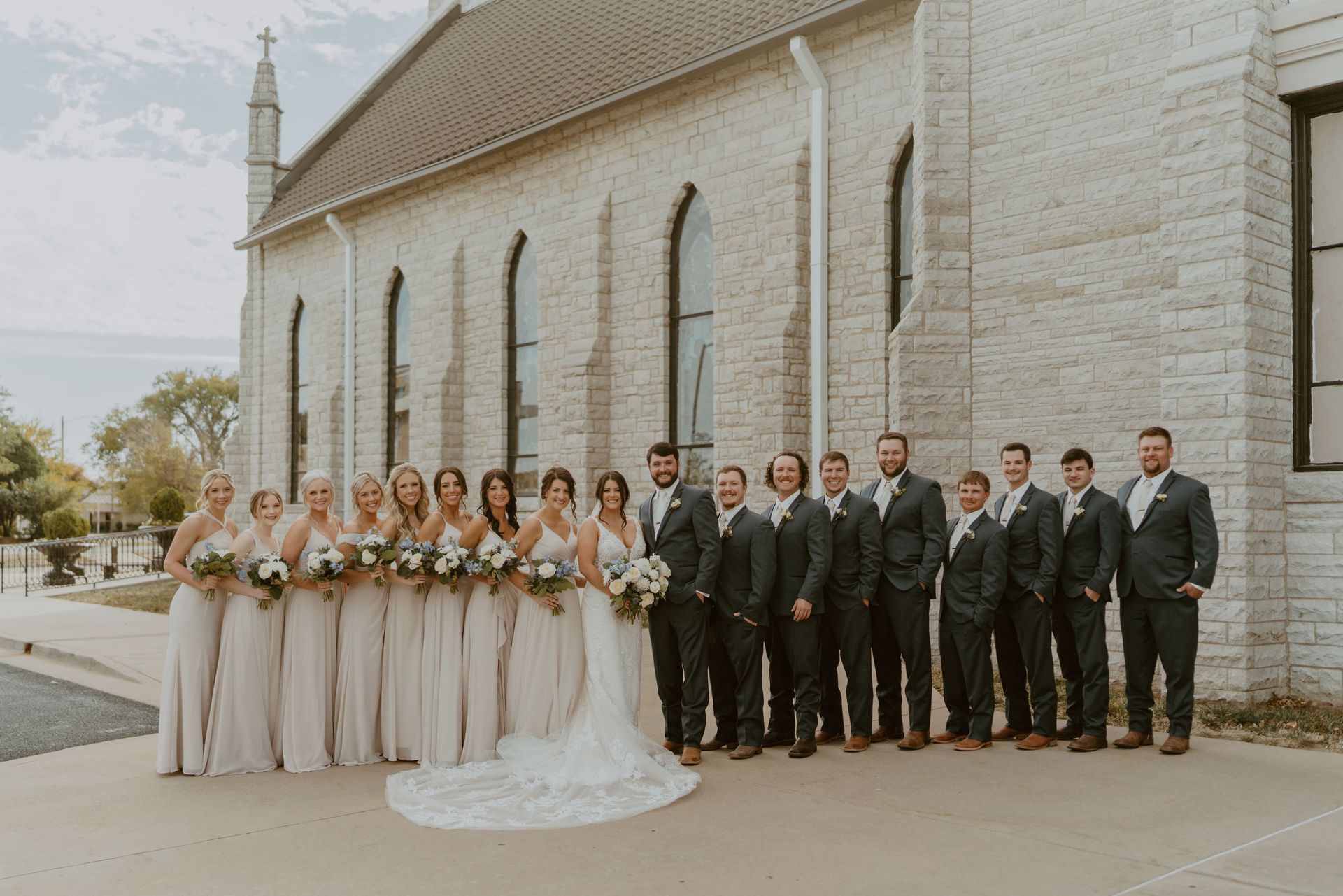 The bride and groom are posing for a picture with their wedding party in front of a church.