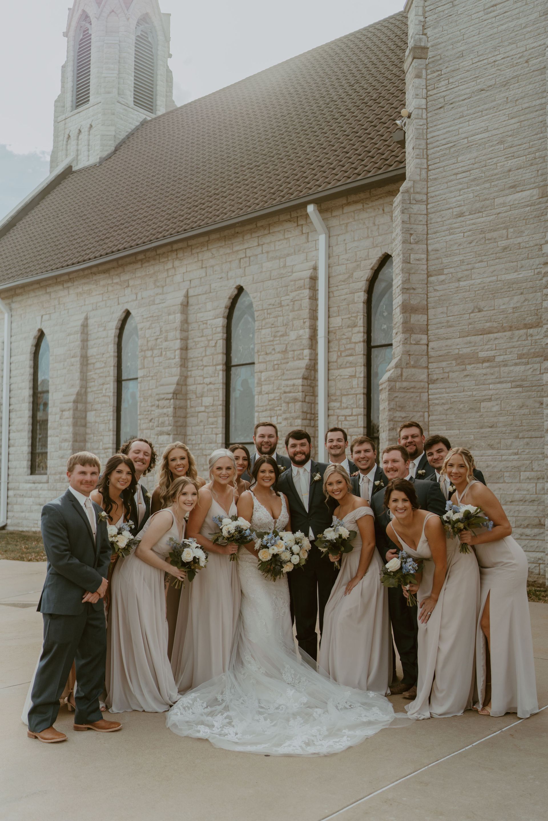 The bride and groom are posing for a picture with their wedding party in front of a church.