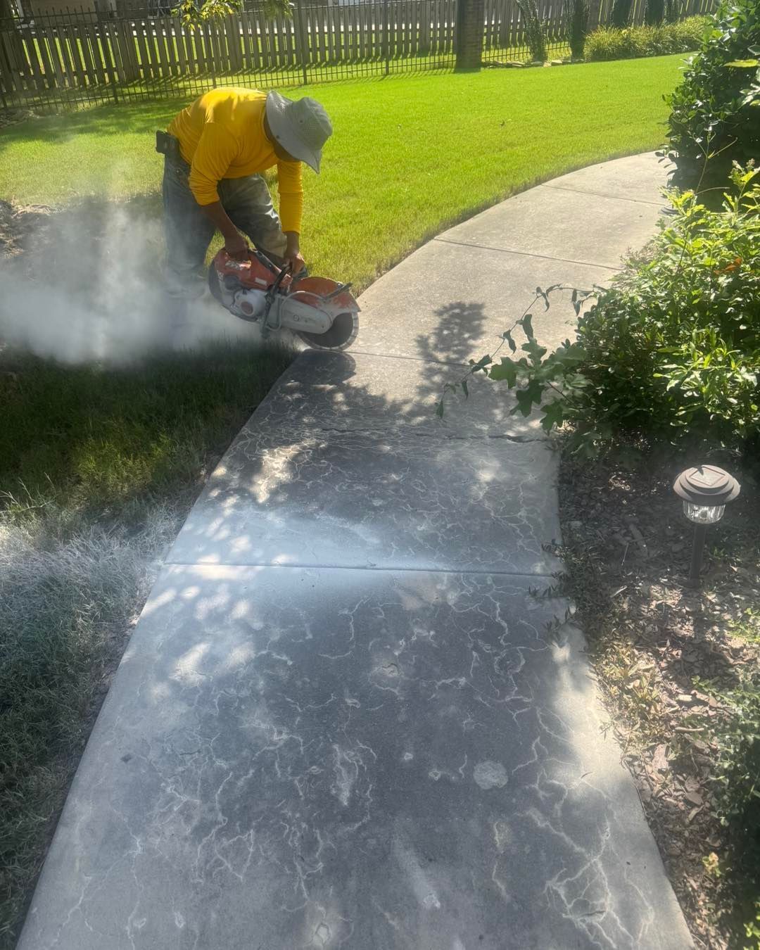Worker cutting concrete sidewalk with a saw, creating dust along a curved path in a sunny yard