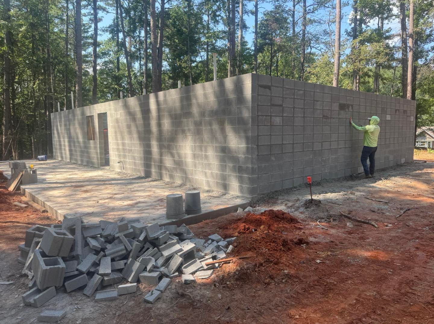Worker building cinder block walls at a forest construction site, with stacked blocks and debris in front.
