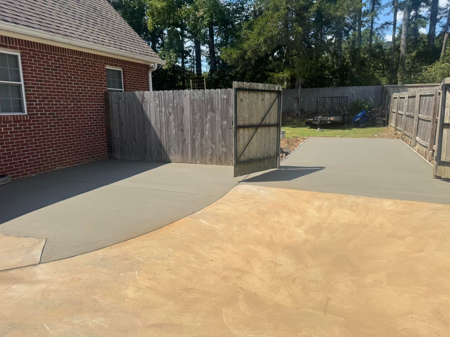 Backyard patio with open wooden gate, concrete area, and tan gravel beside a brick house