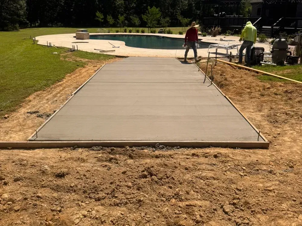 Concrete walkway leading to a backyard swimming pool, with workers and equipment near the pool edge.