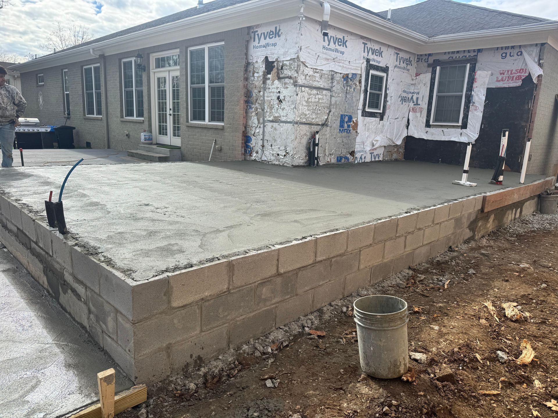 New concrete patio beside a house with unfinished exterior walls and a small bucket in the foreground