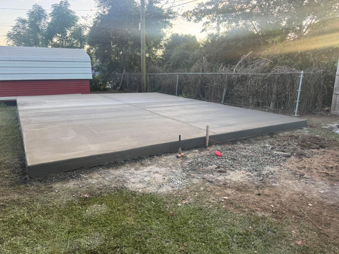 Concrete slab foundation in a backyard beside a red-and-white shed, with trees and fencing in the background