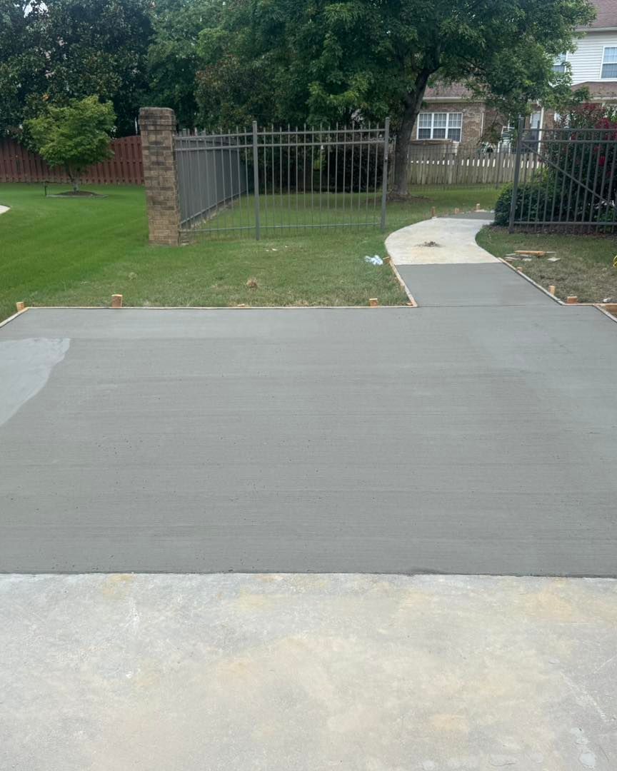 Concrete driveway/path leading to a grassy yard with a black metal fence and trees beyond.