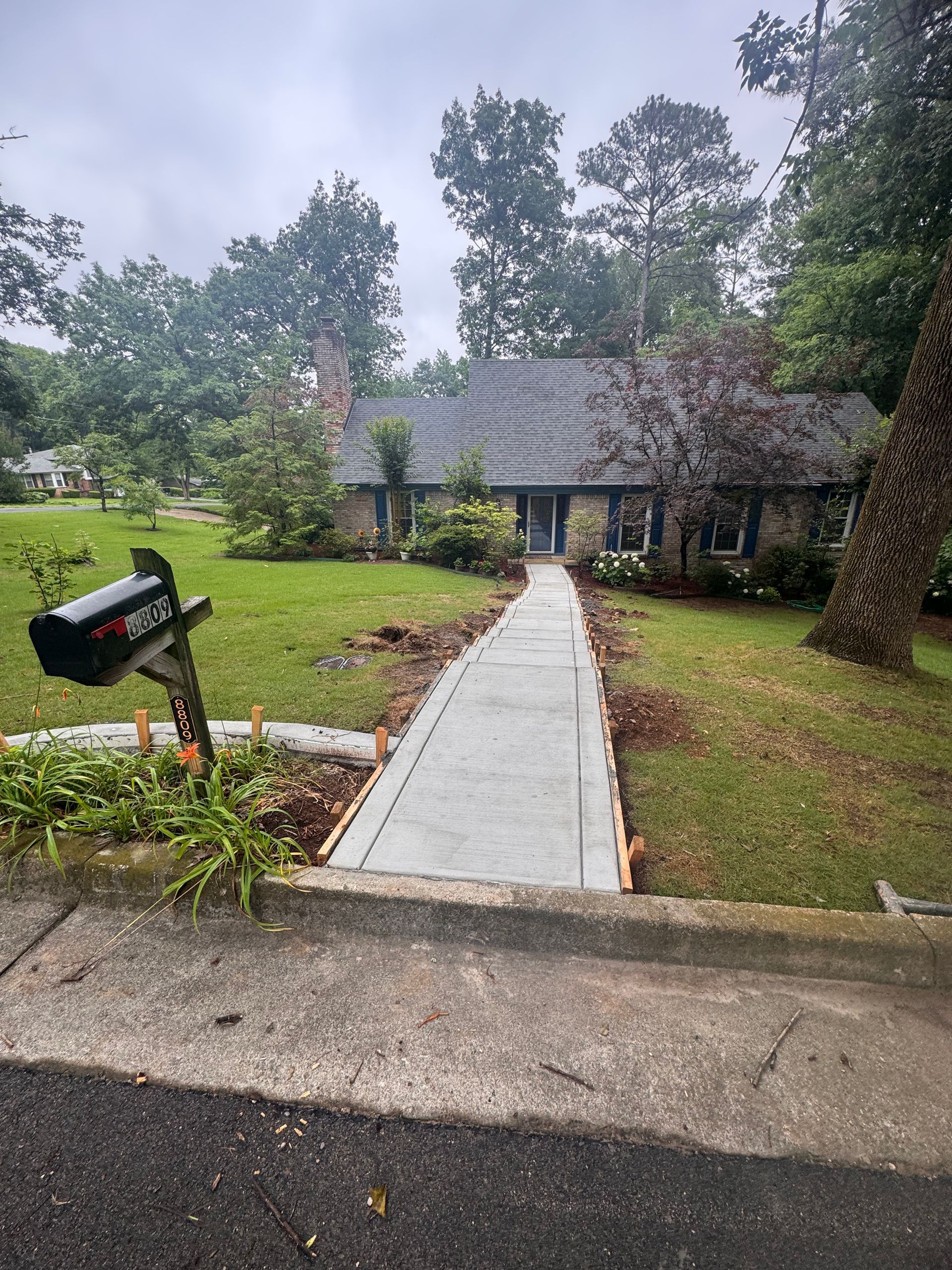 Front walkway leading to a house, with a mailbox and wet lawn on a cloudy day.
