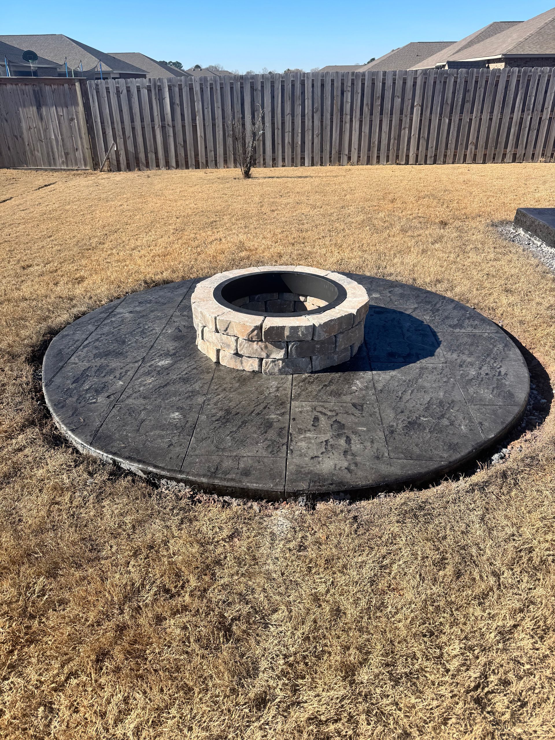 Stone fire pit in a dry backyard with gravel, surrounded by a circular concrete pad and wooden fence.