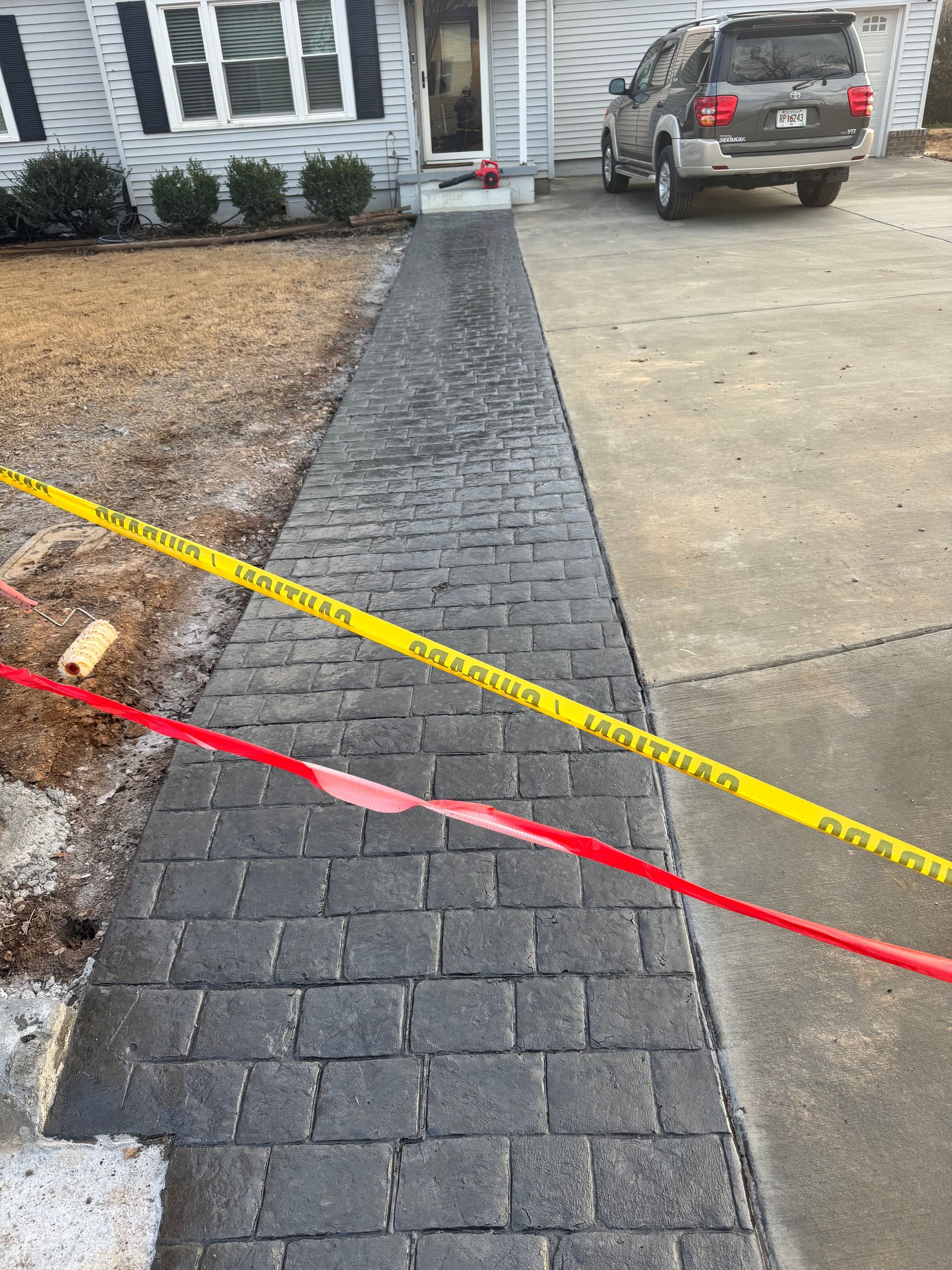 Newly paved brick walkway under construction beside a driveway, marked with red and yellow tape