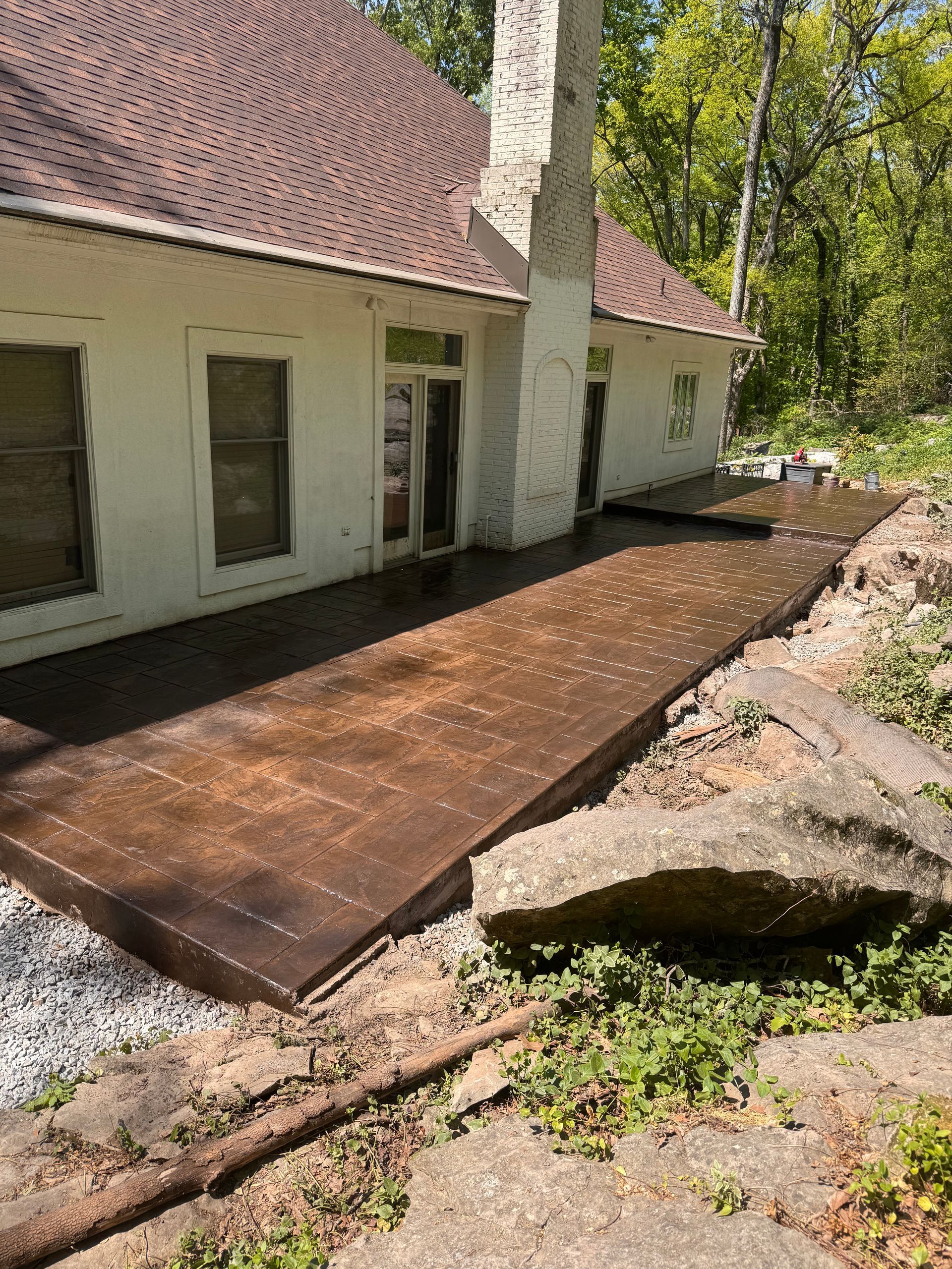 Back patio with dark wood deck beside a white house and red tile roof, bordered by rocks and dirt.